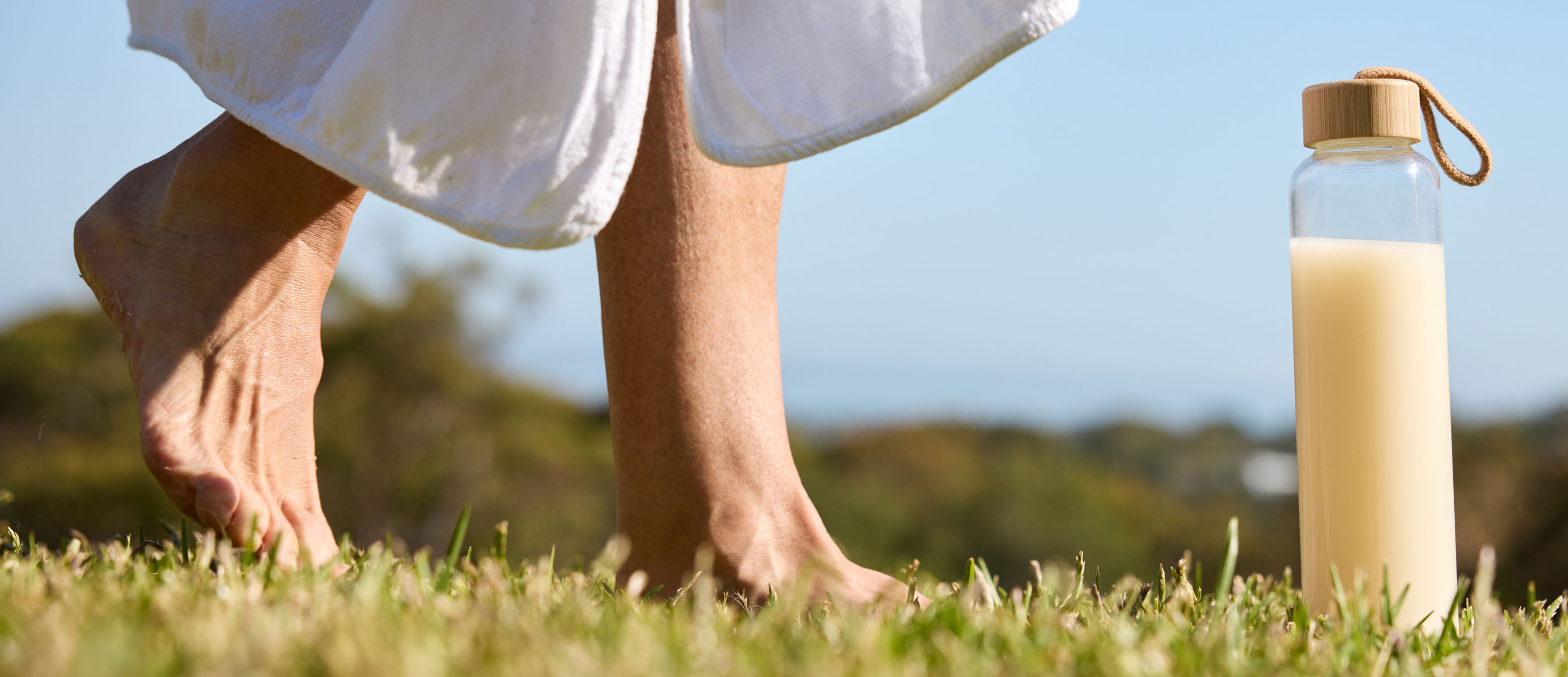 Bare feet grounding on grass beside a glass bottle of SANA protein shake - achieving daily protein goals naturally