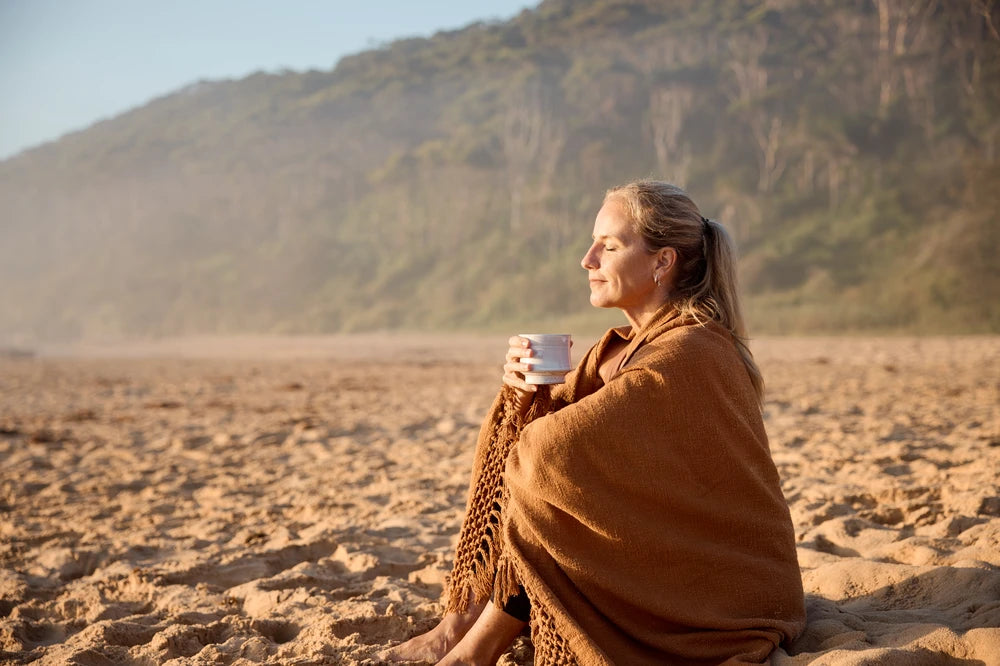 Woman wrapped in a rust-coloured blanket sitting on a sandy beach at sunrise, holding a warm cup and enjoying a mindful morning ritual