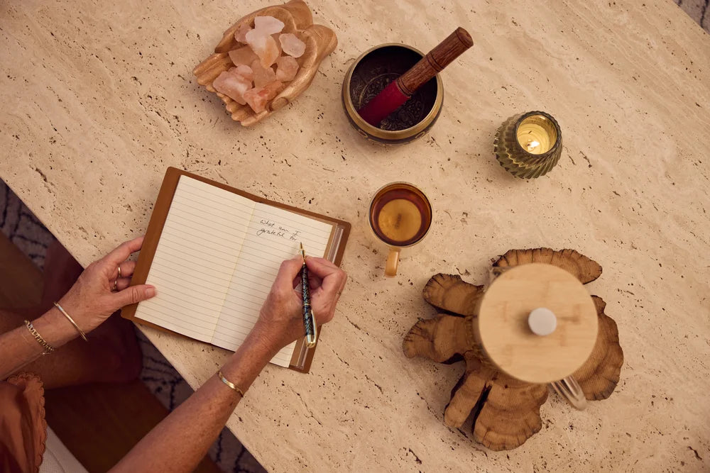 Overhead view of hands writing 'What am I grateful for?' in a journal, surrounded by a singing bowl, rose quartz crystals, herbal tea and a candle on a stone surface