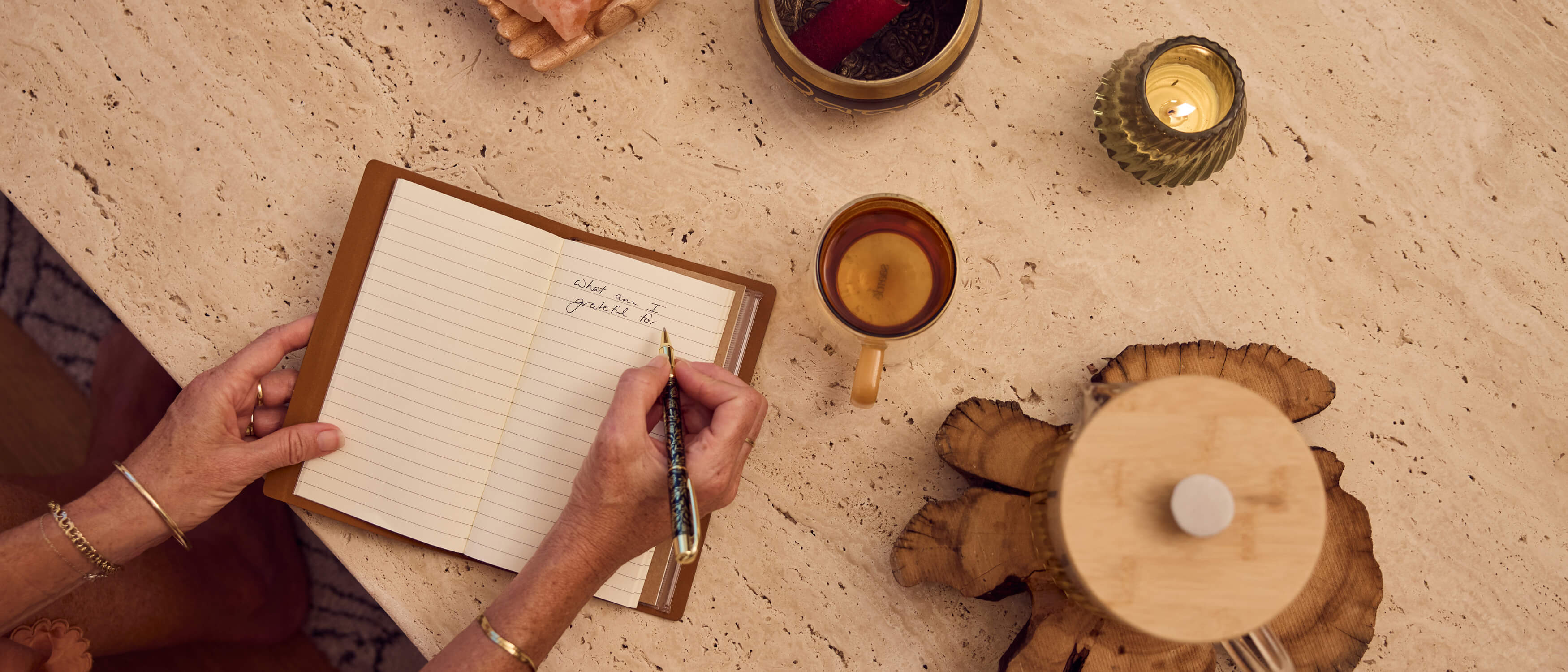 Hands writing gratitude journal with decorative pen surrounded by candles, tea and natural wood elements on travertine surface