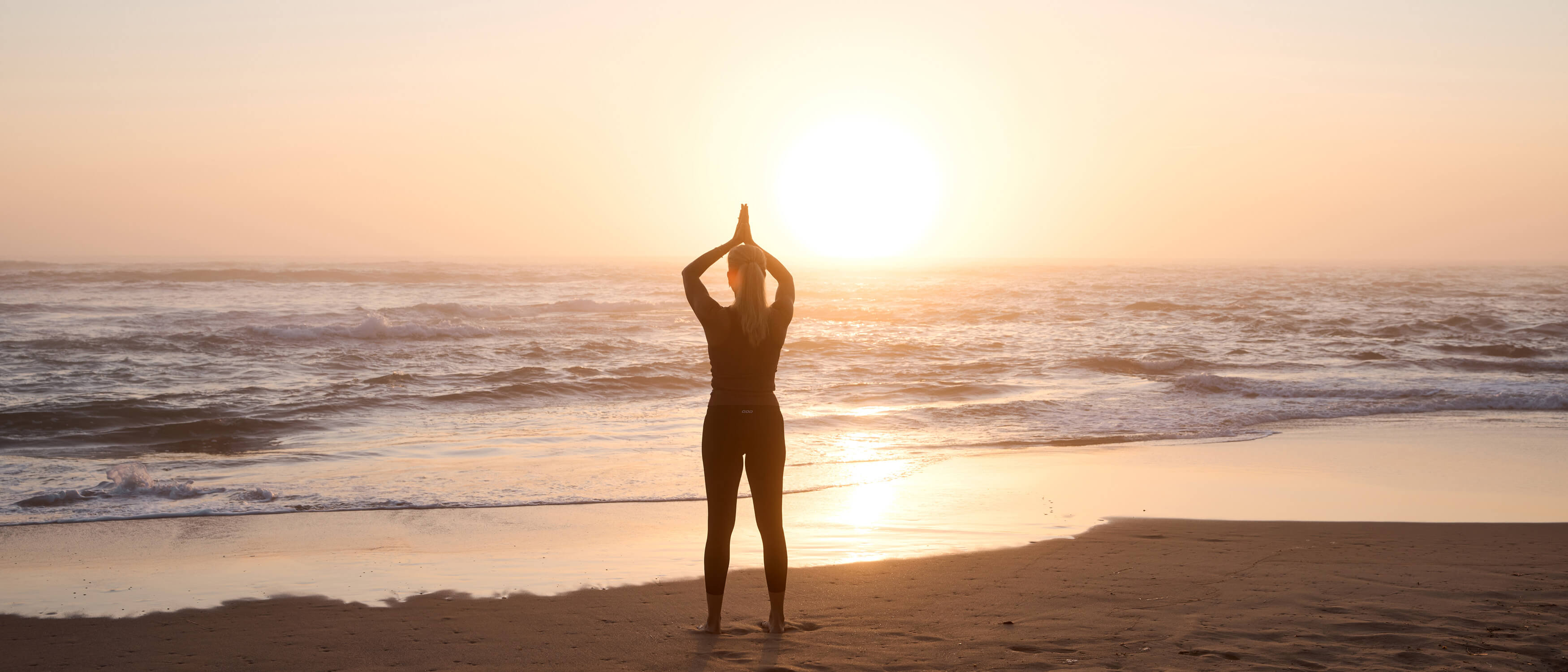 Woman in yoga prayer pose silhouetted against golden sunrise on beach with ocean waves